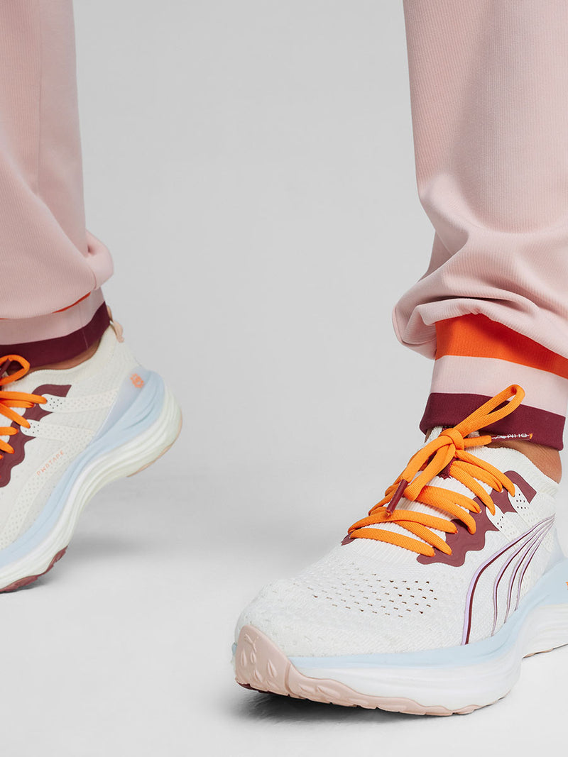 Close up on legs of a Woman Standing Wearing Puma x lemlem Rose Quartz Jumpsuit and Puma x lemlem ForeverRUN Nitro Shoes featuring Icy Blue, Warm White, colors and color details in Cayenne Pepper and Team Regal Red