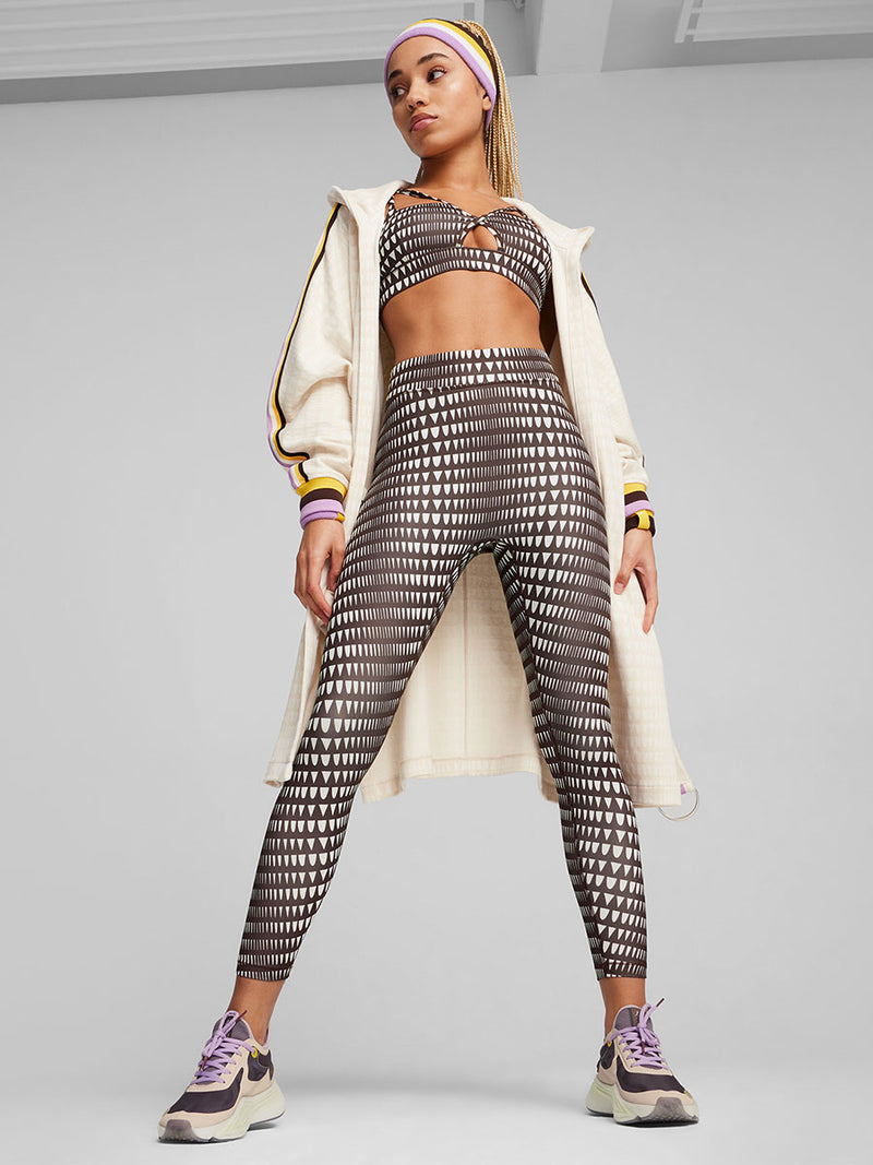 Woman Standing Wearing Puma x lemlem  Low Impact Bra, High Waist Leggings, Anorak , PWR Nitro shoes and  sweatband set featuring color block stripes in yellow, violet, white and dark chocolate colors