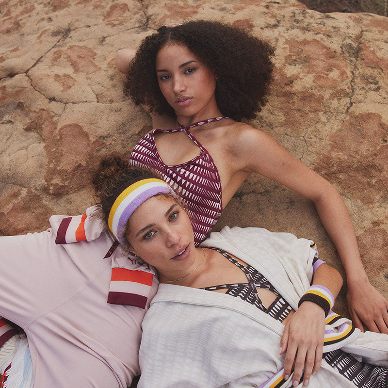 Two woman laying together on a rock. One is wearing matching sport bra and legging in brown and whte and a cream long robe. The other is wearing a light pink fleece jumpsuit tied around a hips and a burgundy leotard under it.