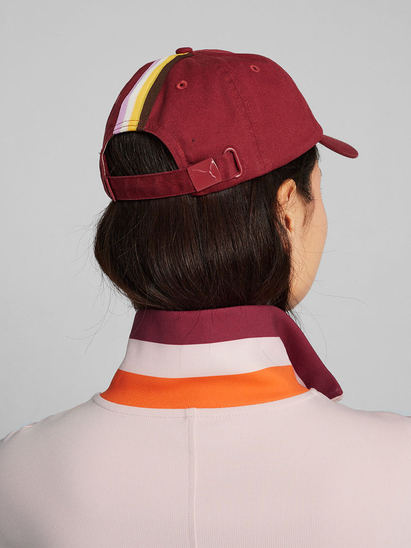 Back View of a Woman Standing Wearing lemlem x Puma Cap featuring Team Regal Red Color and Puma x lemlem Logo on the front and lemlem x Puma jumpsuit