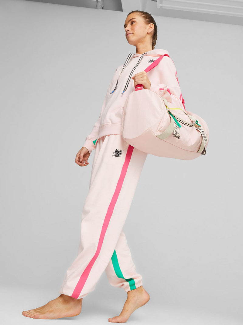 Woman walking wearing Puma x lemlem fleece oversized hoodie and joggers in Frosty Pink Color featuring color pop side stripes in pink and green colors and handsketched puma cat logo and Puma x lemlem studio bag in Frosty Pink Color