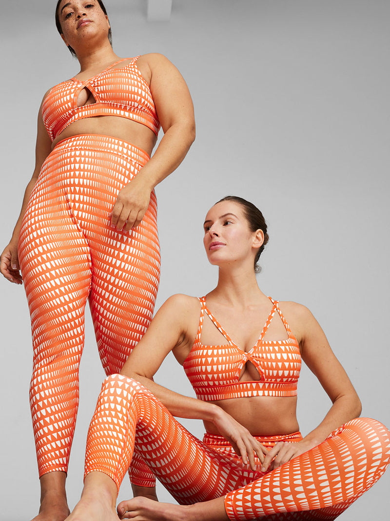 One Woman Standing Wearing Puma x lemlem Low Impact Bra in Cayenne Pepper Color Featuring lemlem Triangle Pattern in White Color and another Woman Sitting Wearing same outfit