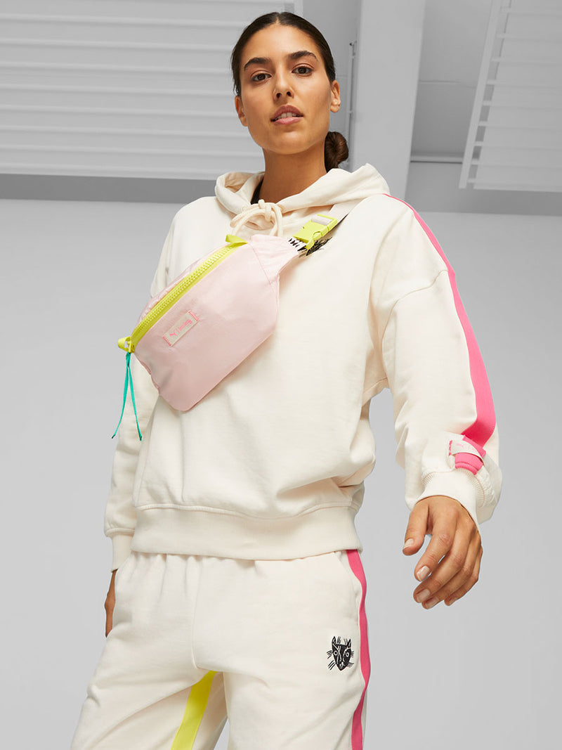 Woman Standing Wearing Puma x lemlem fleece oversized hoodie in ghost pepper color featuring color pop side stripes, Puma x lemlem joggers in ghost pepper color featuring color pop side stripes and hand sketched puma cat logo, and Puma x lemlem waist bag in frosty pink color featuring color block zipper and a shoulder strap with classic lemlem triangle pattern