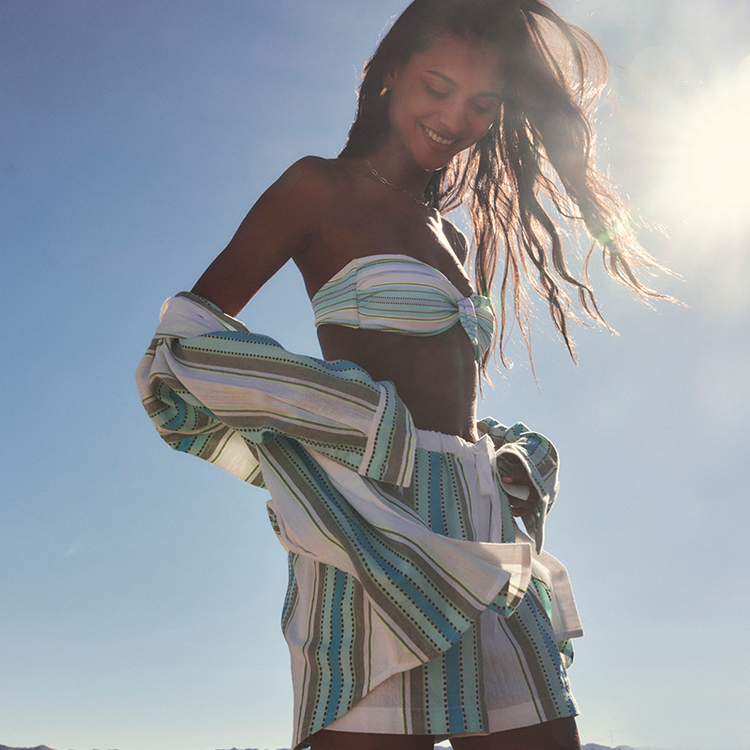 Woman in the sunlight wearing a Genet Mint bikini top with the matching shorts and shirt against a clear blue sky.