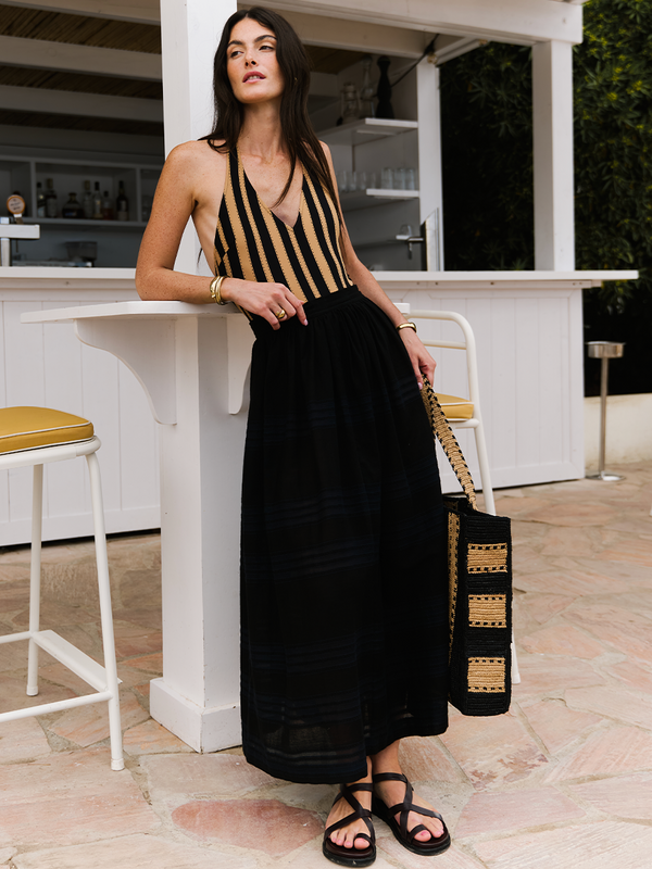 Woman standing by a outdoor bar wearing a lemlem one piece swimsuit with brown and black stripes and a hand woven lemlem black skirt.