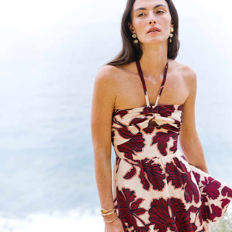 Woman standing in front of the Mediterranean Sea wearing a lemlem linen dress in cream with oversized red floral prints. She is holding a matching pouch. 