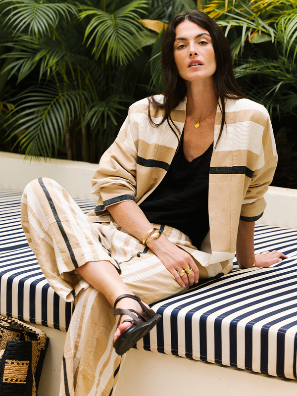 Woman sitting on a stripes outdoor sofa wearing a lemlem handwoven jacket and matching pants in camel.