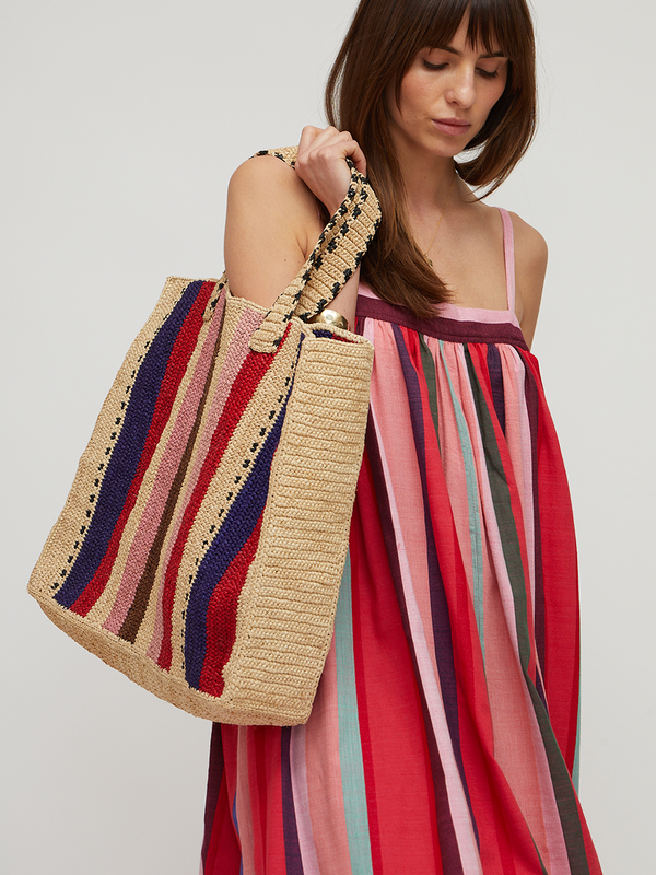 A woman modeling a sleeveless midi dress with vertical pink, red, blue, and teal stripes, paired with matching hand crocheted raffia bag. 