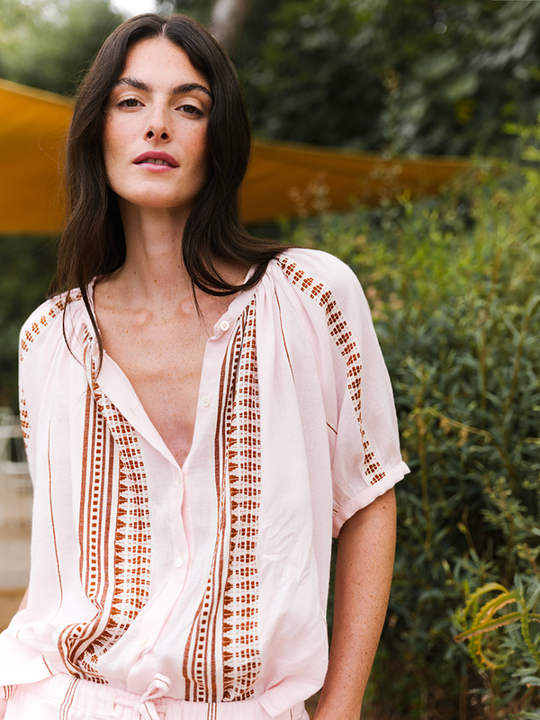 Woman standing in front of trees wearing a lemlem hand woven blouse in light pink with graphic brown tibeb stripes. 