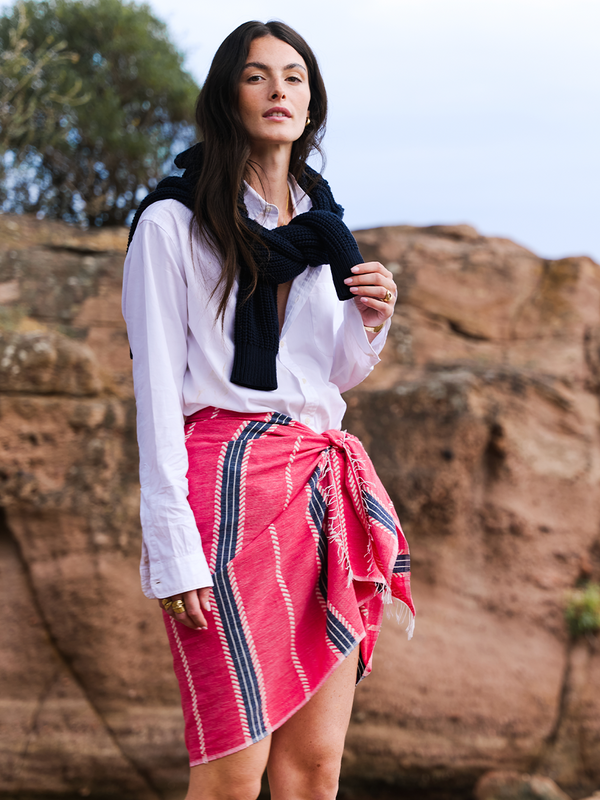 Woman standing by rocky cliff wearing a white shirt with a tied sweater over her shoulder and a lemlem hand woven sarong tied around her waist. 