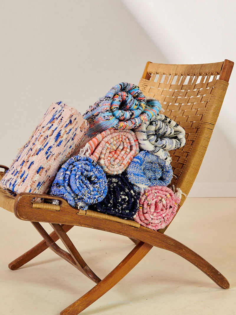 Several multi-colored handwoven rugs, rolled up and stacked in a pile on a wooden lounge chair.