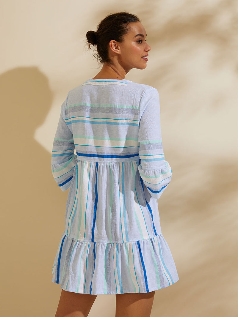 Back view of a woman standing wearing the Ruki Flutter Dress featuring a mutli tonal stripe pattern in five shades of blue with silver and white accents.