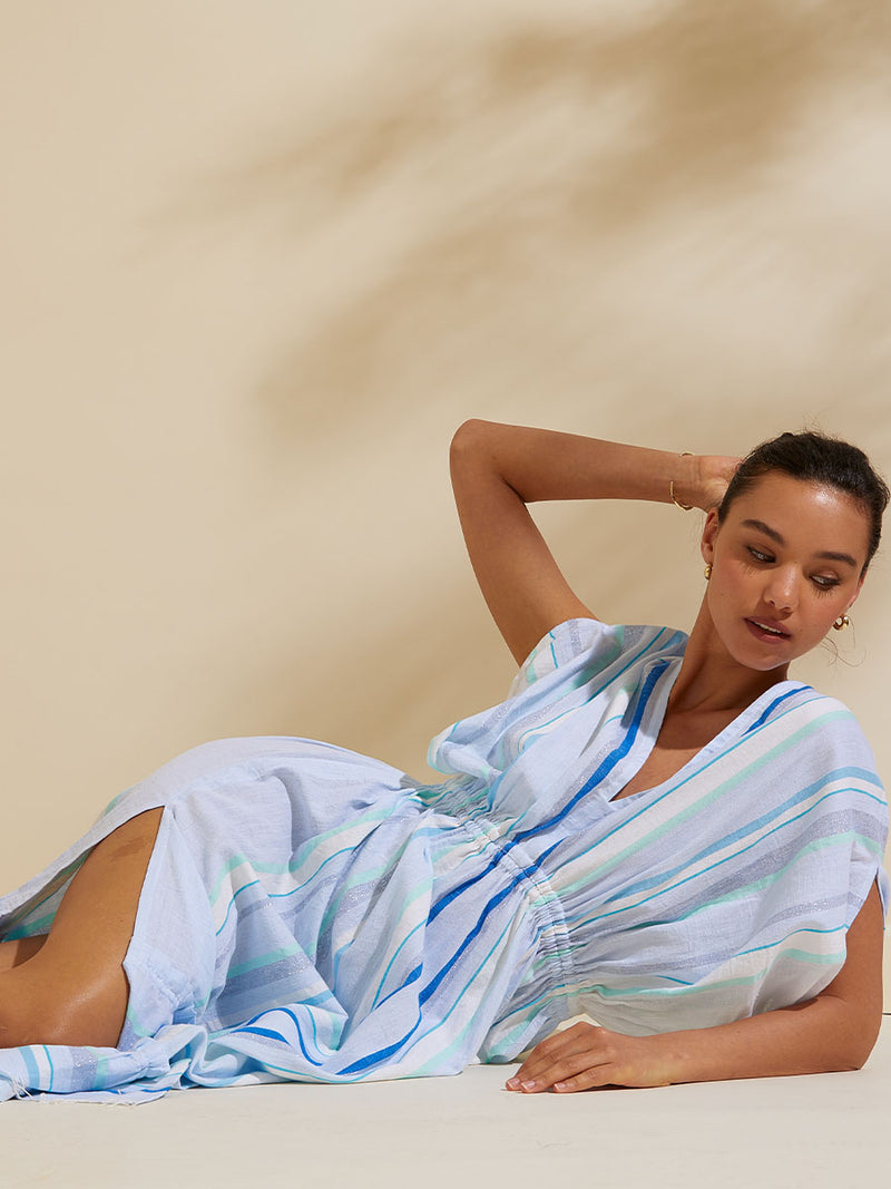 Close up shot of woman laying on the floor on her side wearing the Ruki Plunge Neck Dress featuring a mutli tonal stripe pattern in five shades of blue with silver and white accents.