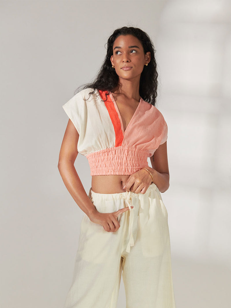 Woman Standing Wearing lemlem Alia Plunge Top Featuring asymmetric color block details in tan and blush colors highlighted with bright orange on the soft cream background.