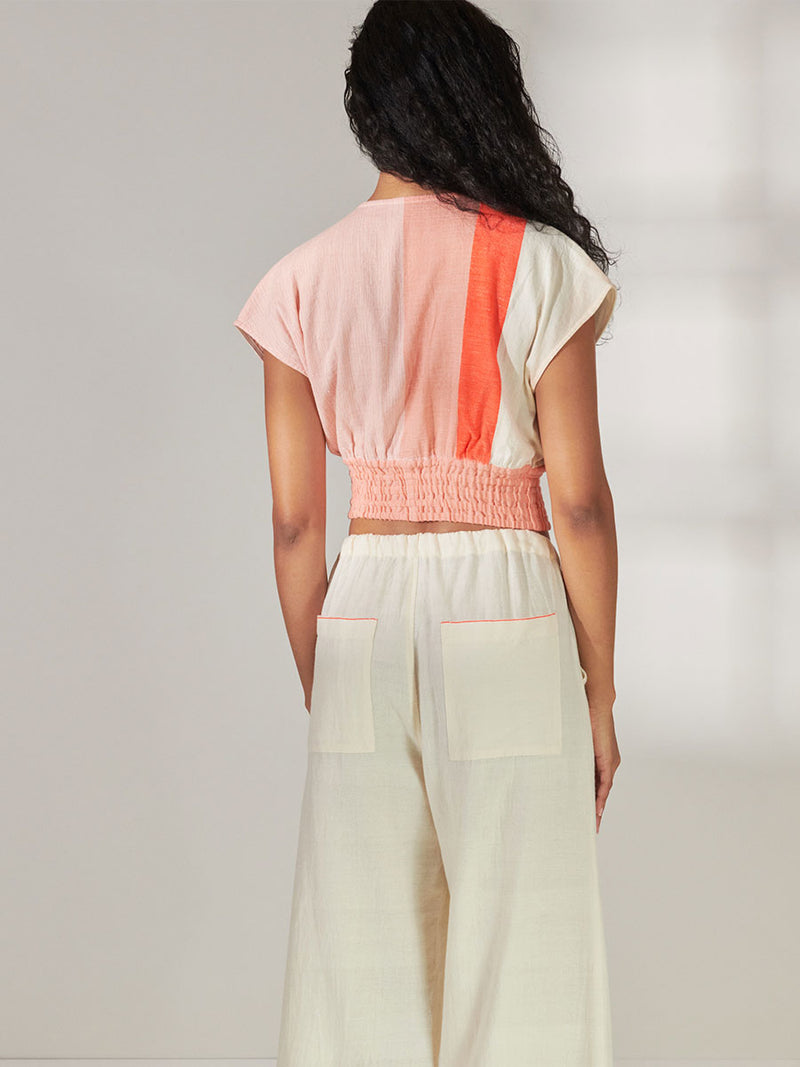 Back View of a Woman Standing Wearing lemlem Alia Plunge Top Featuring asymmetric color block details in tan and blush colors highlighted with bright orange on the soft cream background.