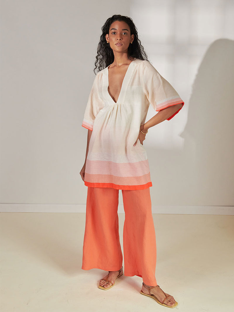 Woman Standing Wearing lemlem Belkis V Neck Caftan Featuring asymmetric color block details in tan and blush colors highlighted with bright orange on the soft cream background and Desta Pants in Kelemi Coral