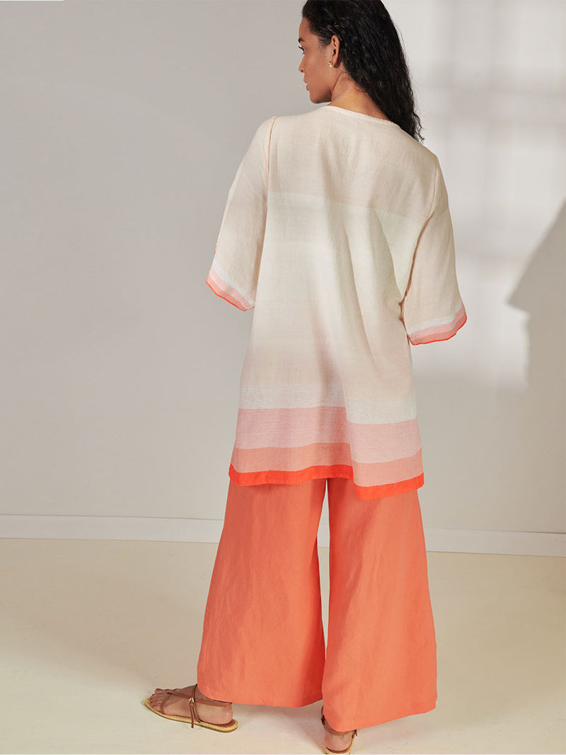 Back View of a Woman Standing Wearing lemlem Belkis V Neck Caftan Featuring asymmetric color block details in tan and blush colors highlighted with bright orange on the soft cream background and Desta Pants in Kelemi Coral