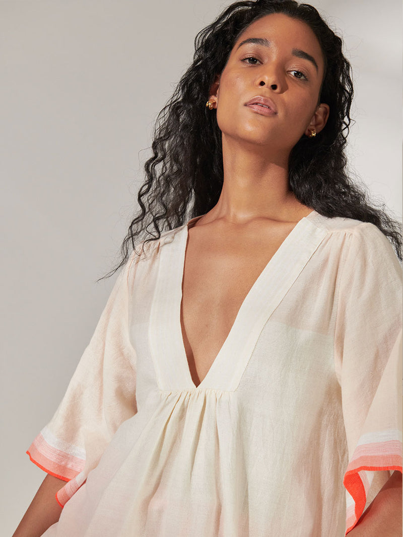 Close up on a Woman Standing Wearing lemlem Belkis V Neck Caftan Featuring asymmetric color block details in tan and blush colors highlighted with bright orange on the soft cream background