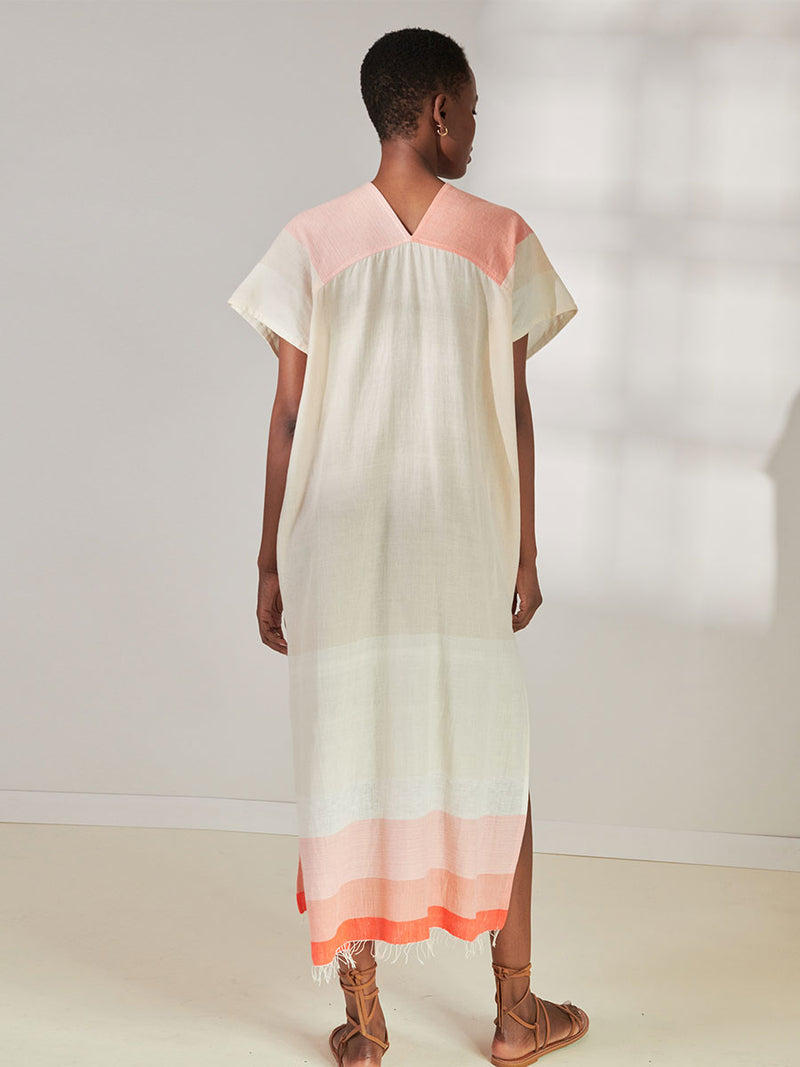 Back View of a Woman Standing Wearing lemlem Gasira V Neck Caftan Featuring asymmetric color block details in tan and blush colors highlighted with bright orange on the soft cream background.