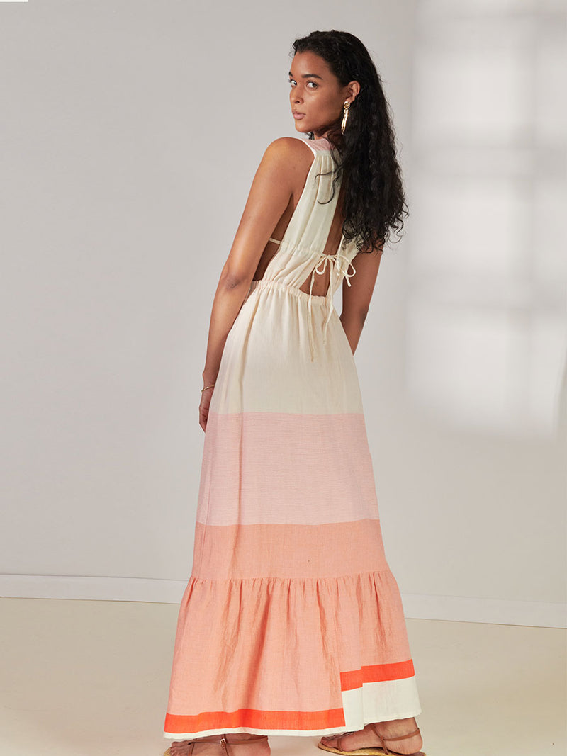 Back view of Woman Standing Wearing lemlem Lelisa V Neck Dress Featuring asymmetric color block details in tan and blush colors highlighted with bright orange on the soft cream background.