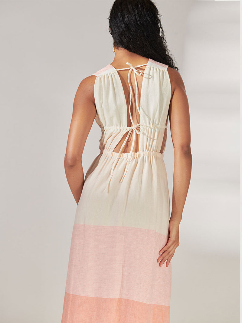 Back view of Woman Standing Wearing lemlem Lelisa V Neck Dress Featuring asymmetric color block details in tan and blush colors highlighted with bright orange on the soft cream background.