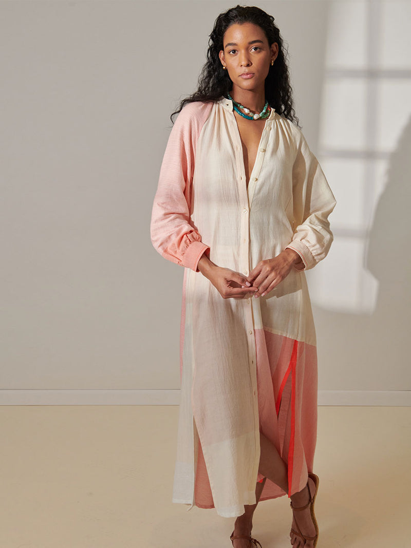 Woman Standing Wearing lemlem Makeda Button Up Dress Featuring asymmetric color block details in tan and blush colors highlighted with bright orange on the soft cream background.