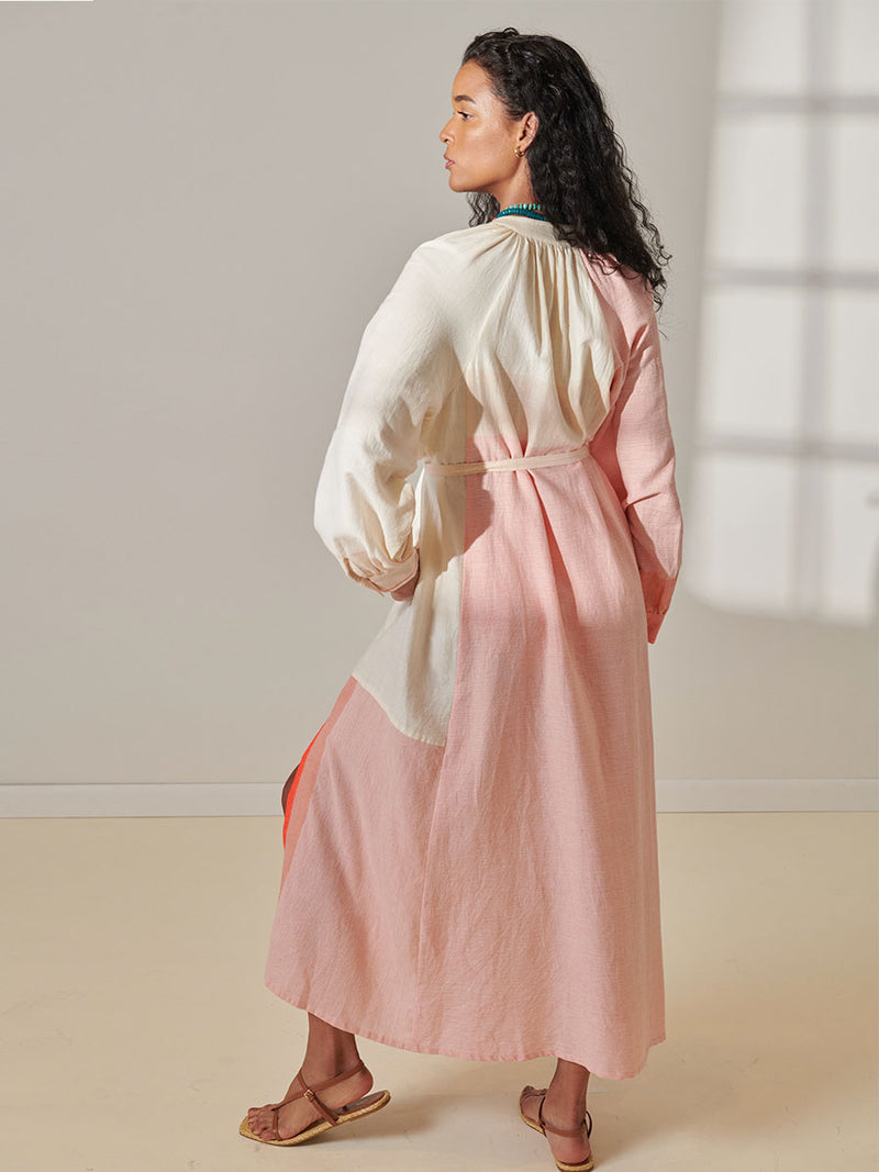 Back View of a Woman Standing Wearing lemlem Makeda Button Up Dress Featuring asymmetric color block details in tan and blush colors highlighted with bright orange on the soft cream background.