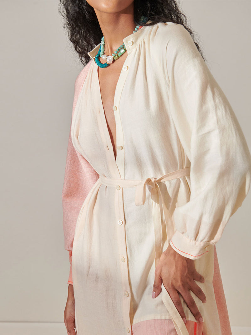 Close up on a Woman Standing Wearing lemlem Makeda Button Up Dress Featuring asymmetric color block details in tan and blush colors highlighted with bright orange on the soft cream background.