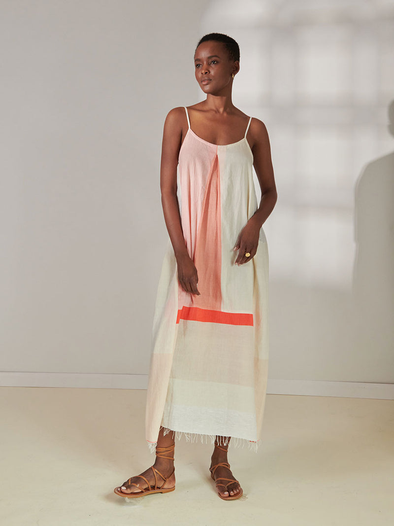 Woman Standing wearing lemlem Nia Slip Dress Featuring asymmetric color block details in tan and blush colors highlighted with bright orange on the soft cream background.
