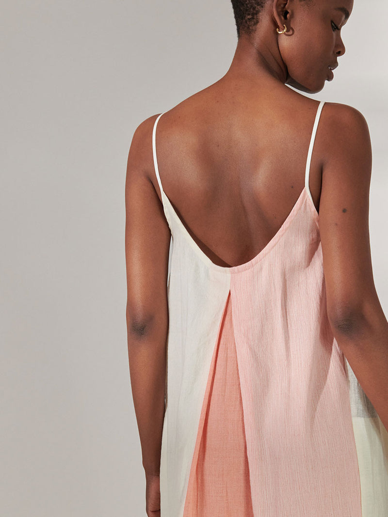 Back View of a Woman Standing wearing lemlem Nia Slip Dress Featuring asymmetric color block details in tan and blush colors highlighted with bright orange on the soft cream background.