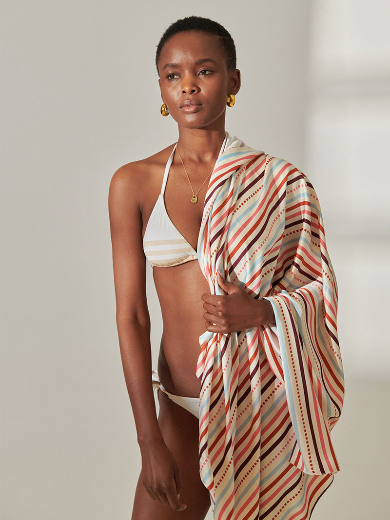 Woman Standing Wearing lemlem Adia Sarong featuring multi stripe pattern with geometric dots with Coral, Sky blue colors grounded by brown ground color on cream background, ayele cappuchino triangle top and string bikini bottom