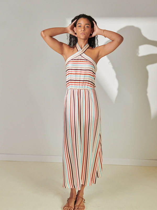Woman Standing Wearing lemlem Ajani Halter Dress featuring multi stripe pattern with geometric dots with Coral, Sky blue colors grounded by brown ground color on cream background.