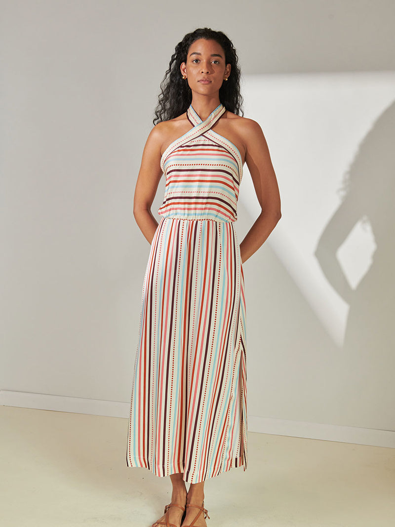 Woman Standing Wearing lemlem Ajani Halter Dress featuring multi stripe pattern with geometric dots with Coral, Sky blue colors grounded by brown ground color on cream background.