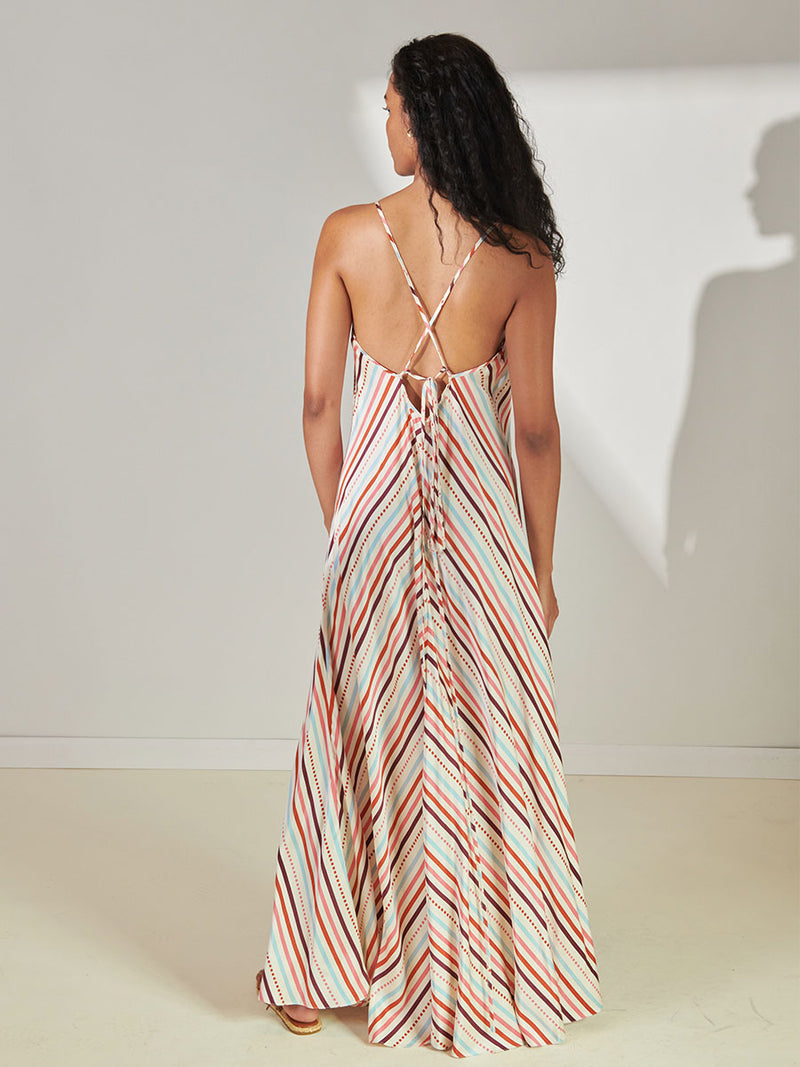 Back View of a Woman Standing Wearing lemlem Aluna Slip Dress featuring multi stripe pattern with geometric dots with Coral, Sky blue colors grounded by brown ground color on cream background.