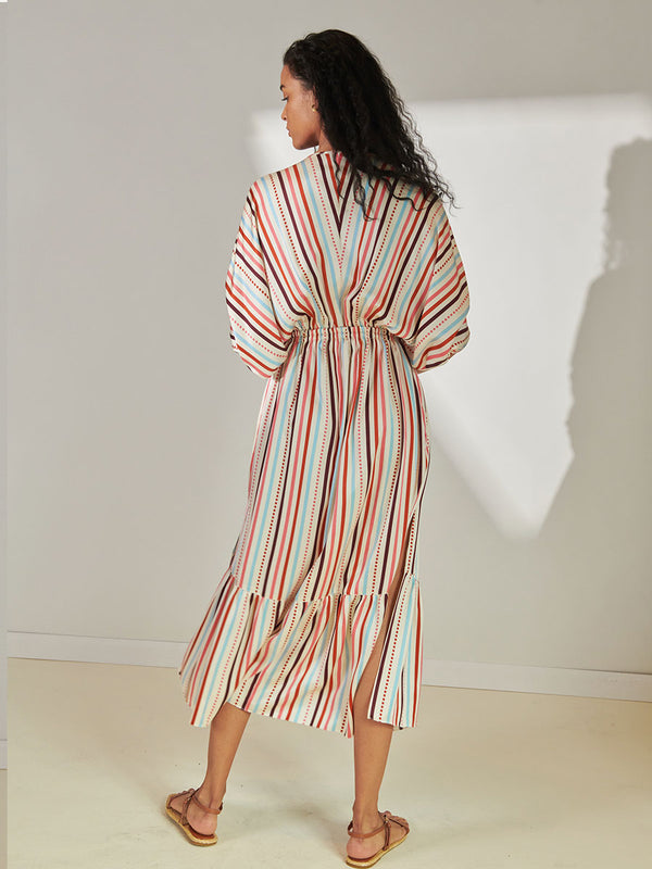 Back View of a Woman Standing Wearing lemlem Leila Plunge Neck Dress featuring multi stripe pattern with geometric dots with Coral, Sky blue colors grounded by brown ground color on cream background.