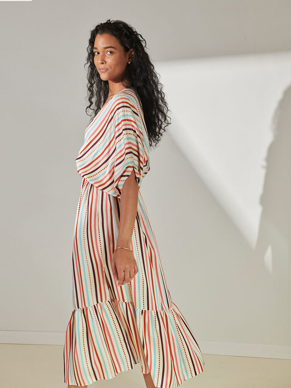 Side View of a Woman Standing Wearing lemlem Leila Plunge Neck Dress featuring multi stripe pattern with geometric dots with Coral, Sky blue colors grounded by brown ground color on cream background.