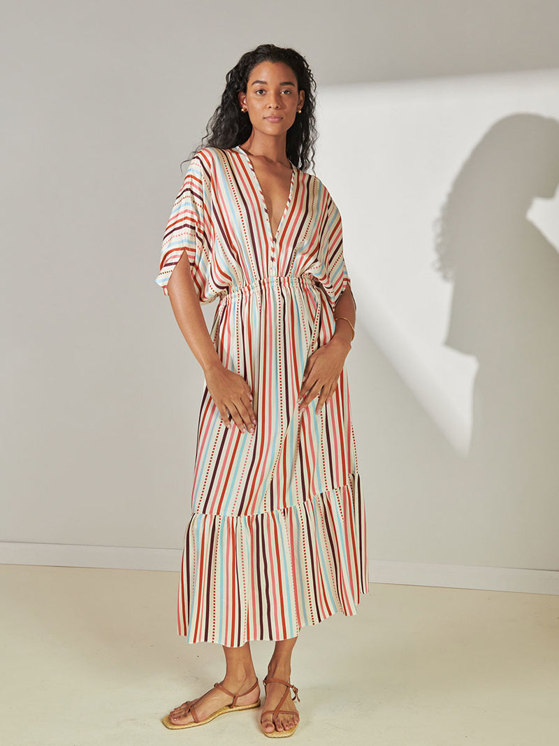 Woman Standing Wearing lemlem Leila Plunge Neck Dress featuring multi stripe pattern with geometric dots with Coral, Sky blue colors grounded by brown ground color on cream background.