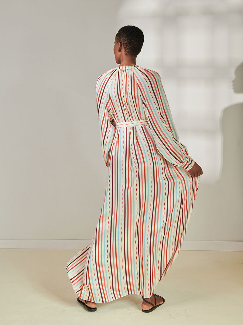 Back View of a Woman Standing Wearing lemlem Makeda Button Up Dress featuring multi stripe pattern with geometric dots with Coral, Sky blue colors grounded by brown ground color on cream background.