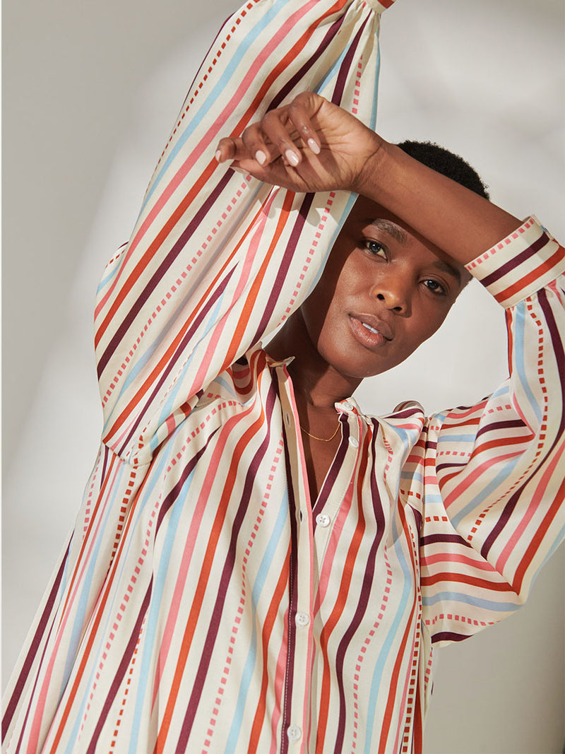 Close up on a Woman Standing Wearing lemlem Makeda Button Up Dress featuring multi stripe pattern with geometric dots with Coral, Sky blue colors grounded by brown ground color on cream background.