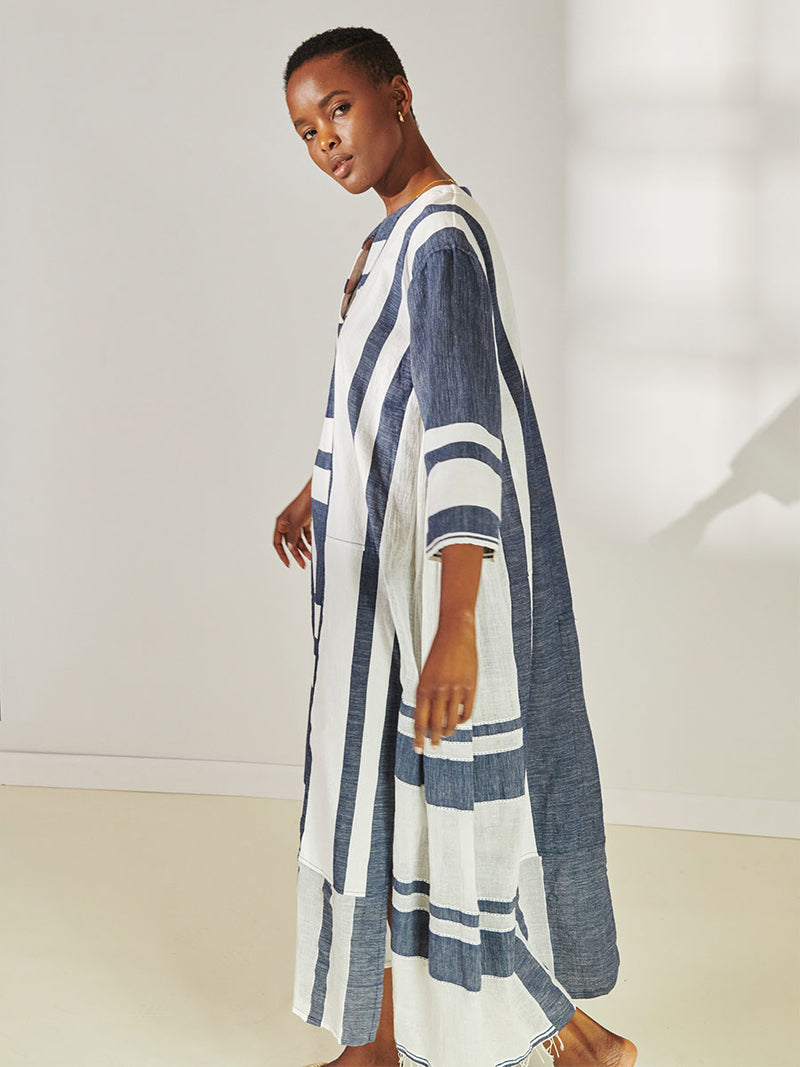 Side View of a Woman Standing Wearing lemlem Fana Caftan Featuring Bold Stripe Pattern with pick stitch edge in Classic Navy and White colors.
