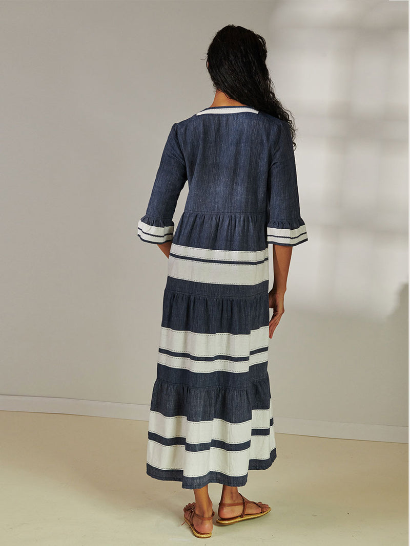 Back View of a Woman Standing Wearing lemlem Hawi Flutter Dress Featuring Bold Stripe Pattern with pick stitch edge in Classic Navy and White colors.