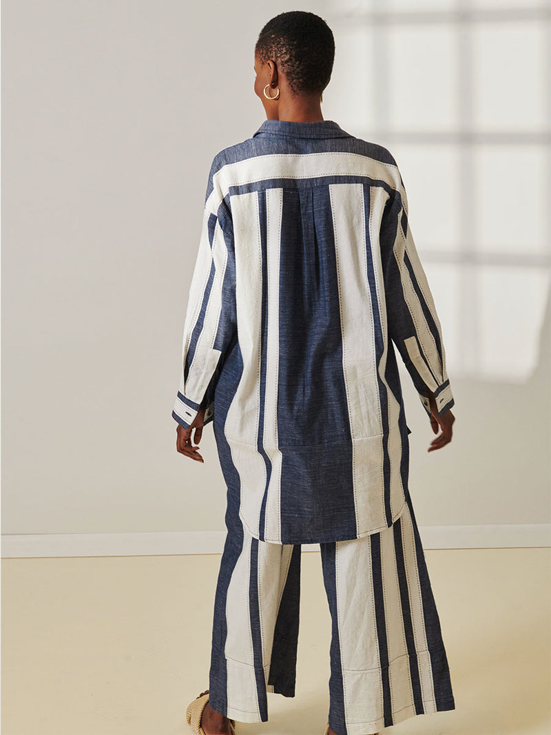 Back View of a Woman Standing Wearing lemlem Mariam Shirt Featuring Bold Stripe Pattern with pick stitch edge in Classic Navy and White colors and matching Desta Pants