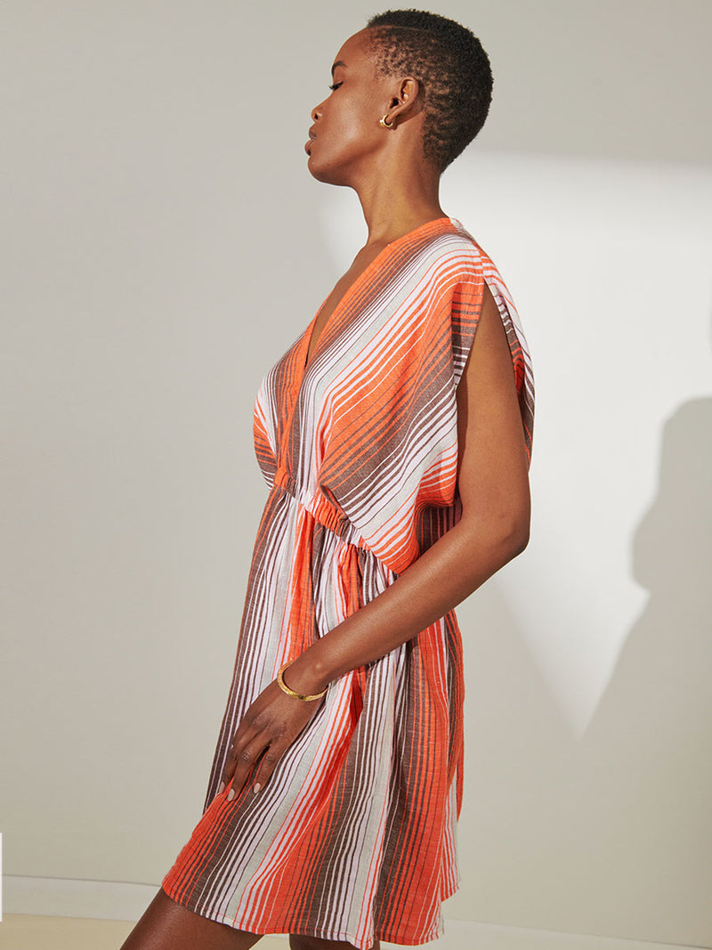 Side View of a Woman Standing Wearing lemlem Alem Short Plunge Dress featuring graded continuous stripe pattern creating an ombre effect featuring earth, orchid & burnt orange.