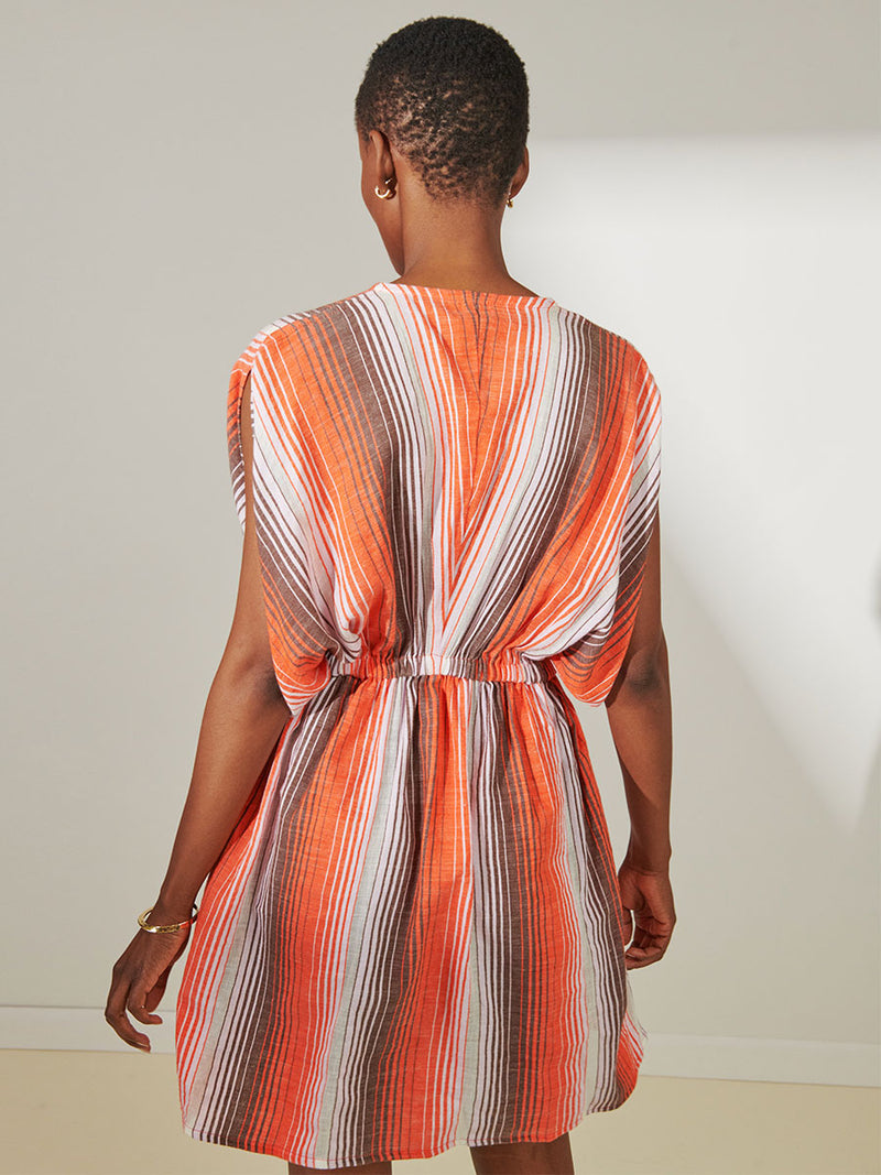 Back View of a Woman Standing Wearing lemlem Alem Short Plunge Dress featuring graded continuous stripe pattern creating an ombre effect featuring earth, orchid & burnt orange.