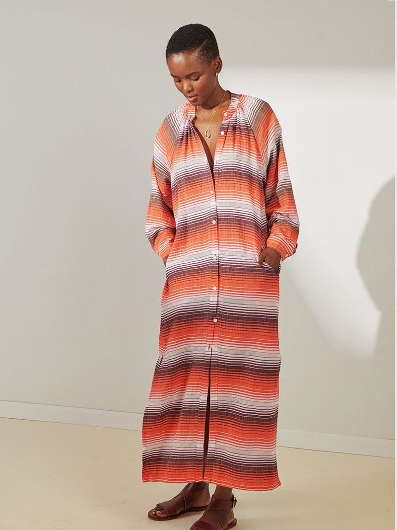 Woman Standing Wearing lemlem Makeda Button Up Dress featuring graded continuous stripe pattern creating an ombre effect featuring earth, orchid & burnt orange.