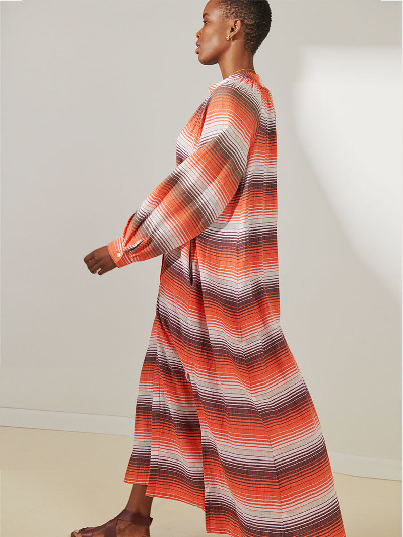 Side View of a Woman Standing Wearing lemlem Makeda Button Up Dress featuring graded continuous stripe pattern creating an ombre effect featuring earth, orchid & burnt orange.