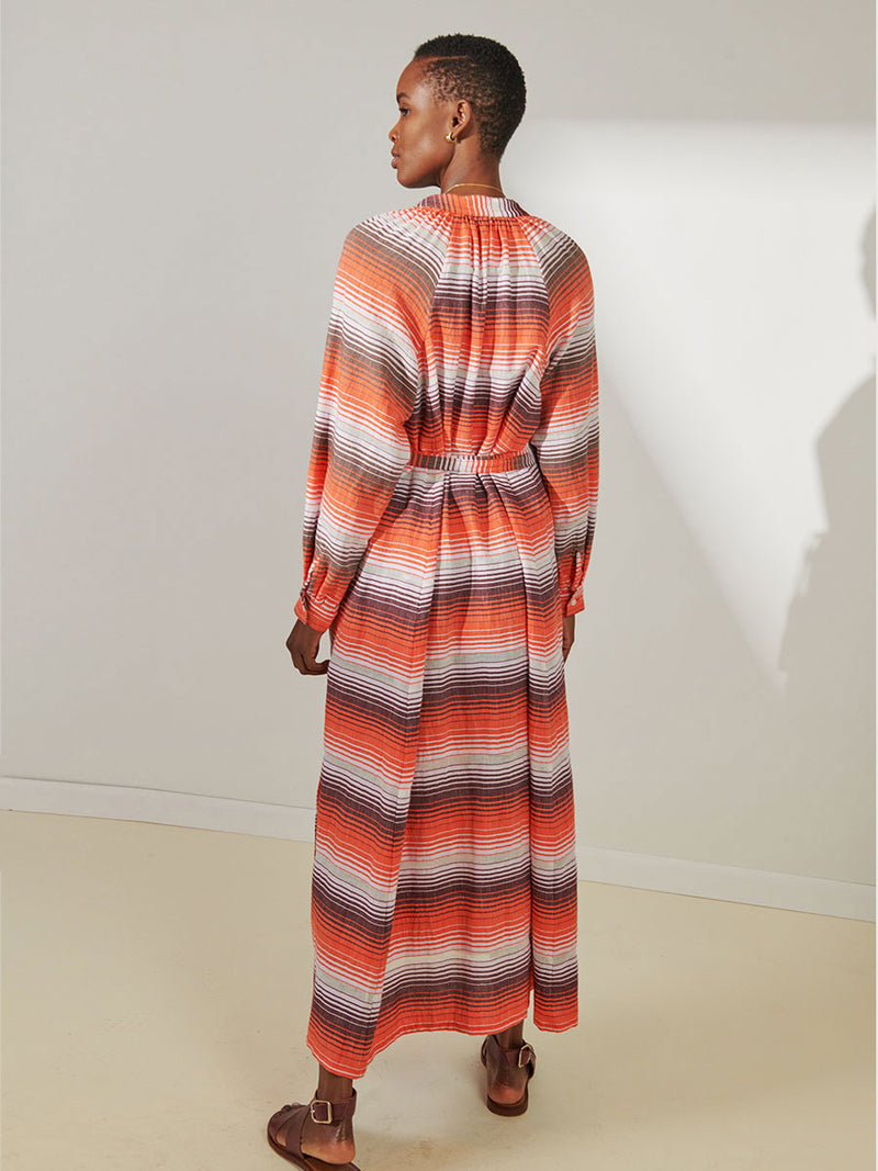 Back View of a Woman Standing Wearing lemlem Makeda Button Up Dress featuring graded continuous stripe pattern creating an ombre effect featuring earth, orchid & burnt orange.