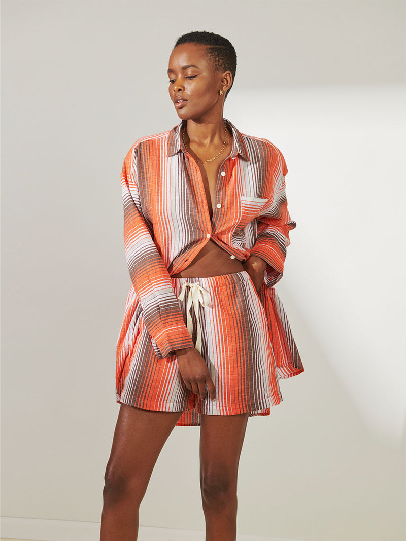 Woman Standing Wearing lemlem Mariam Shirt featuring graded continuous stripe pattern creating an ombre effect featuring earth, orchid & burnt orange and matching shorts