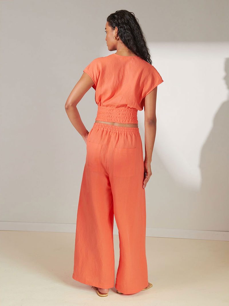Back View of a Woman Standing Wearing lemlem Alia Plunge Top in Coral Color and Desta Pants in coral color