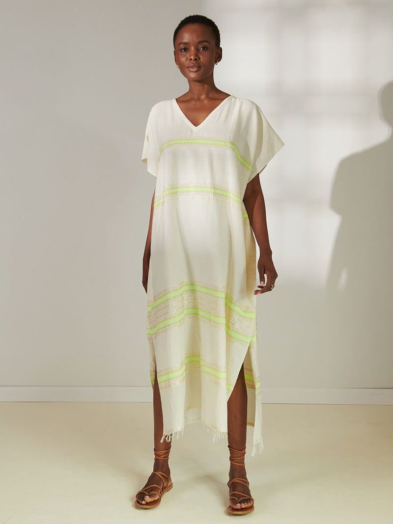 Woman Standing Wearing lemlem Dalila Caftan featuring combination of matte and shine natural tibebs and stripes in Vanilla Cream and Lime sorbet colors.